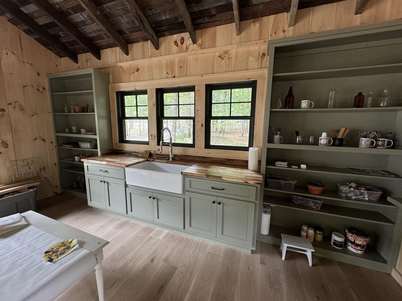 Photo of rustic cabin with open shelving on either side of a kitchen sink. The dark wood exposed ceilings, exposed wood walls and hardwood flooring give the cabin a rustic feel