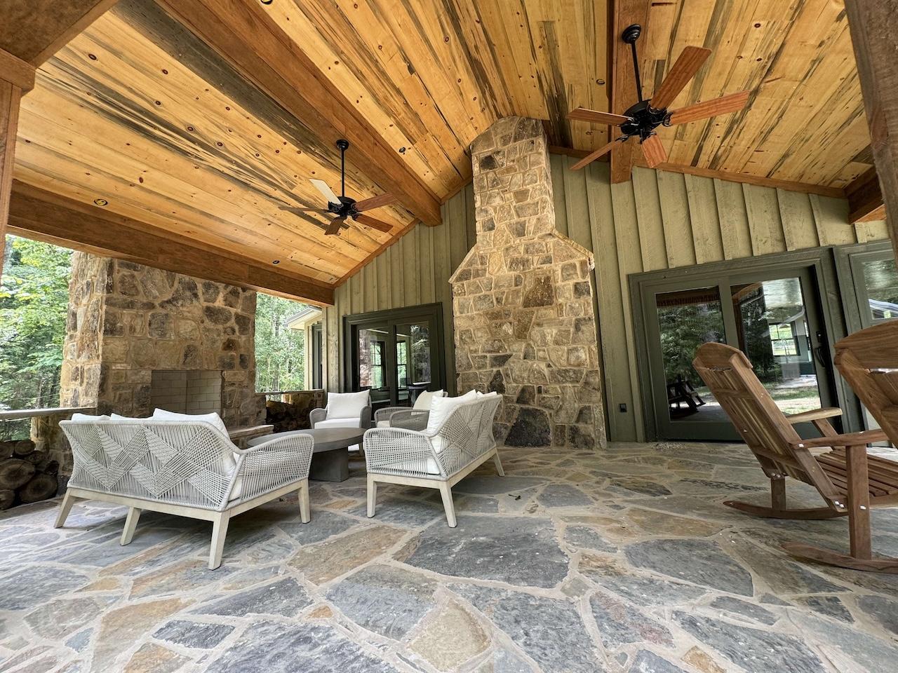 Photo of porch with exposed wood ceiling with two ceiling fans covering a stone patio flooring and a stone fireplace on the exterior wall of the porch