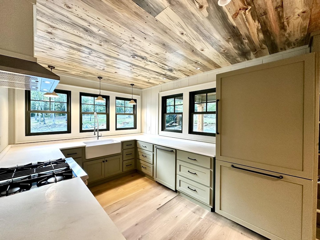 Photo of rustic cabin with open shelving on either side of a kitchen sink. The dark wood exposed ceilings, exposed wood walls and hardwood flooring give the cabin a rustic feel