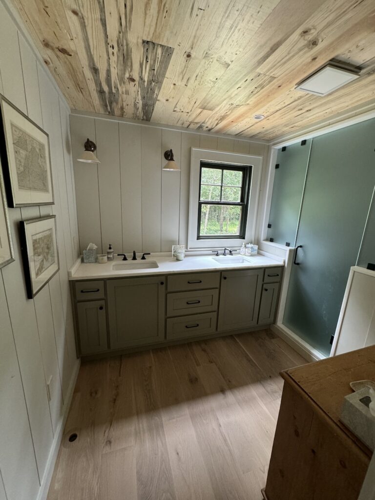 Photo of rustic cabin with open shelving on either side of a kitchen sink. The dark wood exposed ceilings, exposed wood walls and hardwood flooring give the cabin a rustic feel