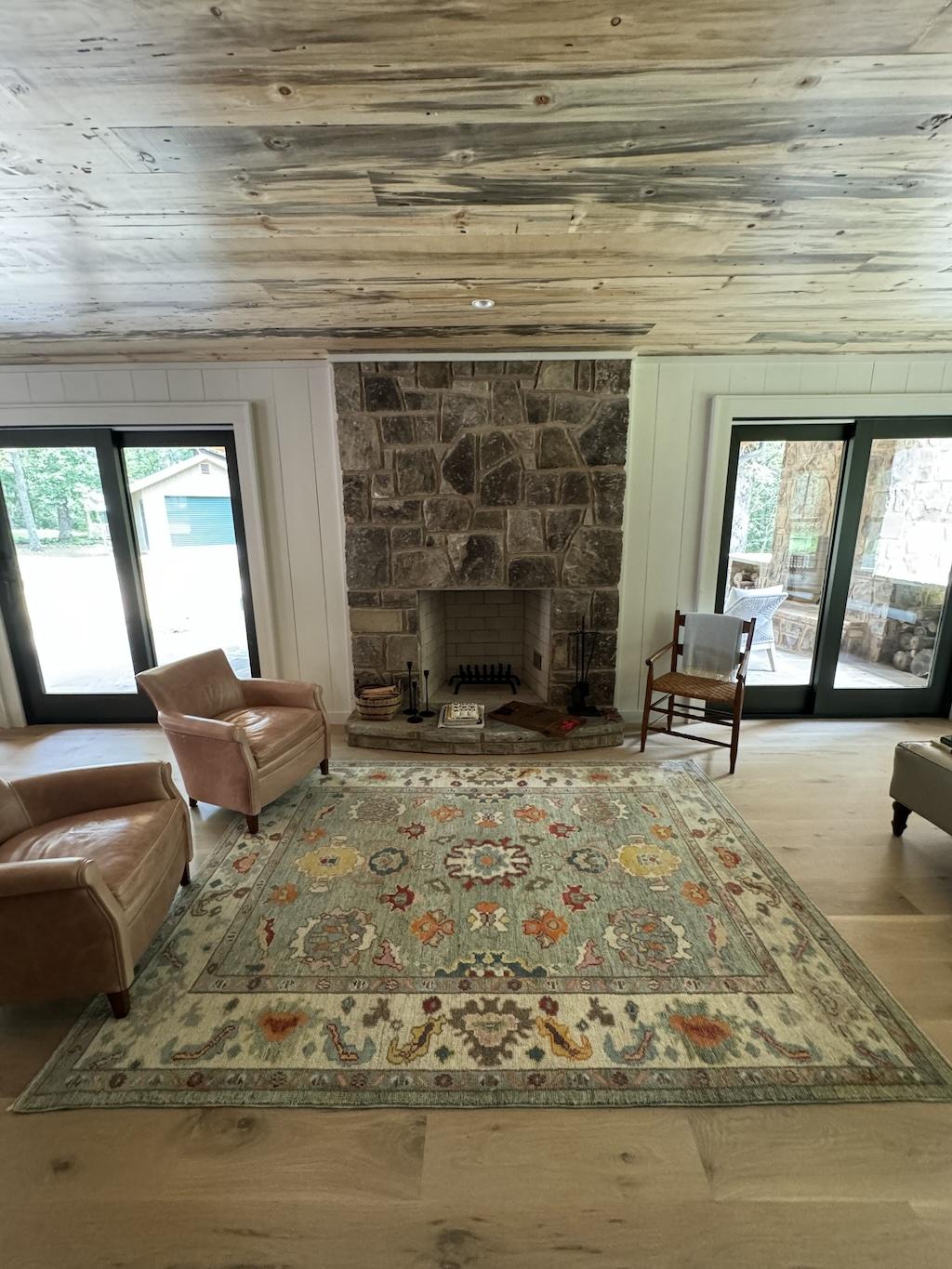 Photo of living room with exposed wood ceiling and hardwood floors. Glass doors are on either side of the stone fireplace