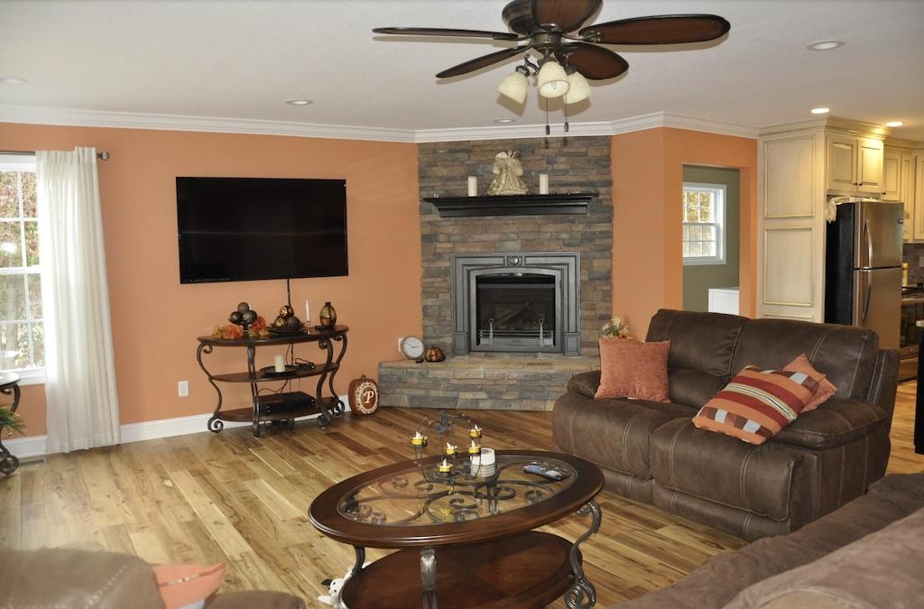 Photo of living room with built in corner fireplace with brick facade. High polish hardwood wide plank floors and an open kitchen can been seen in photo